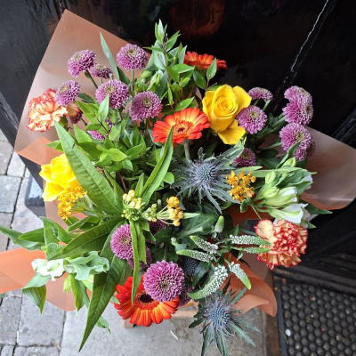 Vibrant Summer Hand-Tie - A colourful celebration of yellow roses, orange and red gerbera, purple chrysanthemums, blue thistle and ruffled carnations with fresh foliage. Bright, bold and full of life.