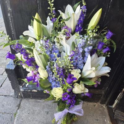 Midnight Blue Sympathy Bouquet - A serene hand-tie of pure white lilies, blue and purple delphinium, campanula and stocks, tied with a soft lilac ribbon. A calm, dignified arrangement, perfect for sympathy or a peaceful gift.